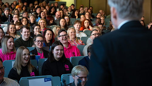 Im Vordergrund steht ein Mann mit grauen Haaren im Anzug, der von hinten zu sehen ist. Vor ihm sitzen auf den Reihen eines Hörsaals viele Studierende, die vergnügt lachen.
